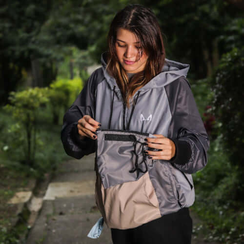 Woman wearing a gray and beige dog training windbreaker holding the removable insulated pouch with a logo in a forest setting