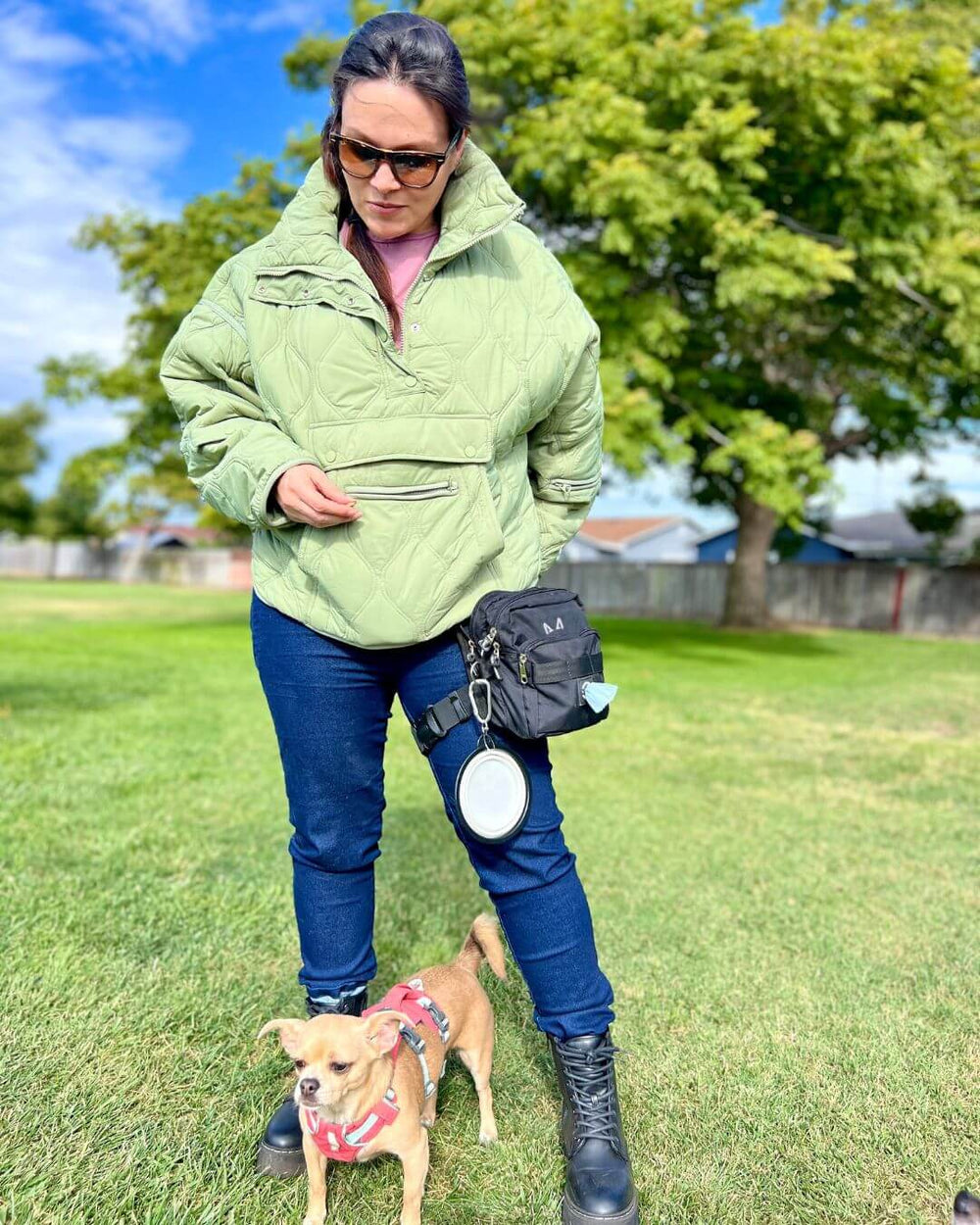Woman wearing the thigh dog treat bag in a green jacket training a small dog in a park