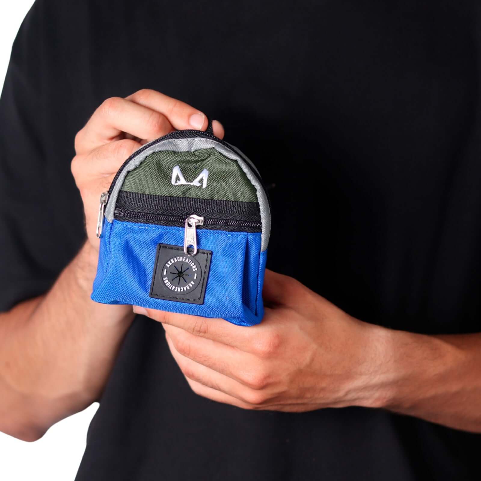 Person holding a small blue and green poop bag and treat holder with a brand logo against a black background